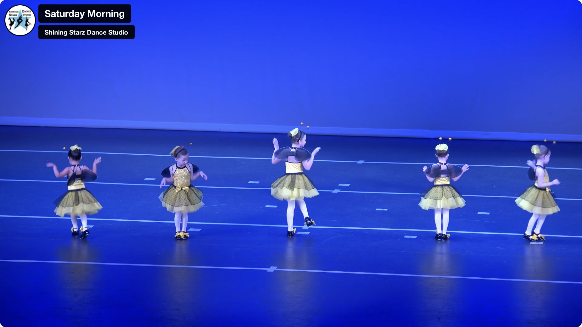 Two children in bee-themed costumes perform a dance routine on stage under blue lighting as part of the Recital Package for Two Students, moving with raised or extended arms and facing different directions.